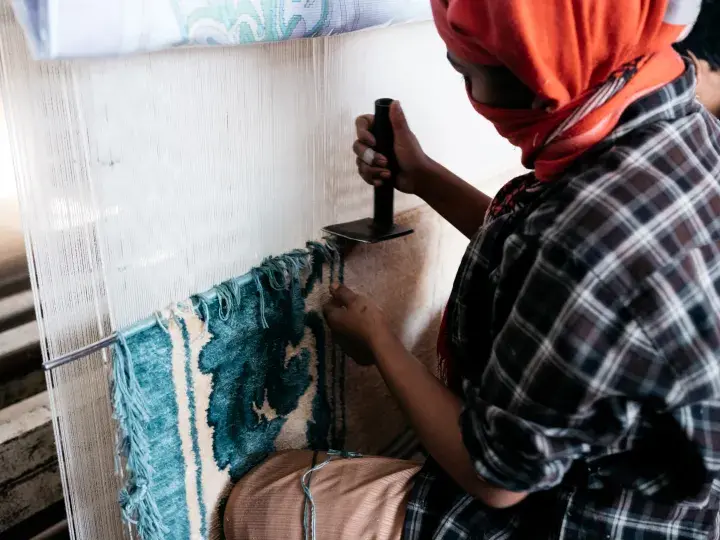 Person with an orange head scarf works on a white and blue rug, Rugs 368 © The Studio M – thestudiom.com, courtesy of GoodWeave International